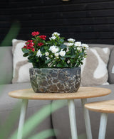 Une Plante Artificielle Patio Rose 35 cm crème UV se dresse sur une table ronde en bois devant un canapé beige avec des coussins à motifs.