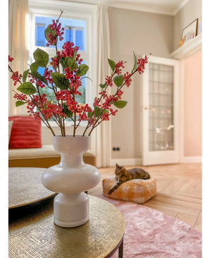 Sur une table se trouve un vase blanc avec des baies Kunstttak roses (64 cm) et des feuilles vertes. En arrière-plan, un chat est allongé sur un coussin rose dans un salon lumineux aux murs beiges et à la porte vitrée.