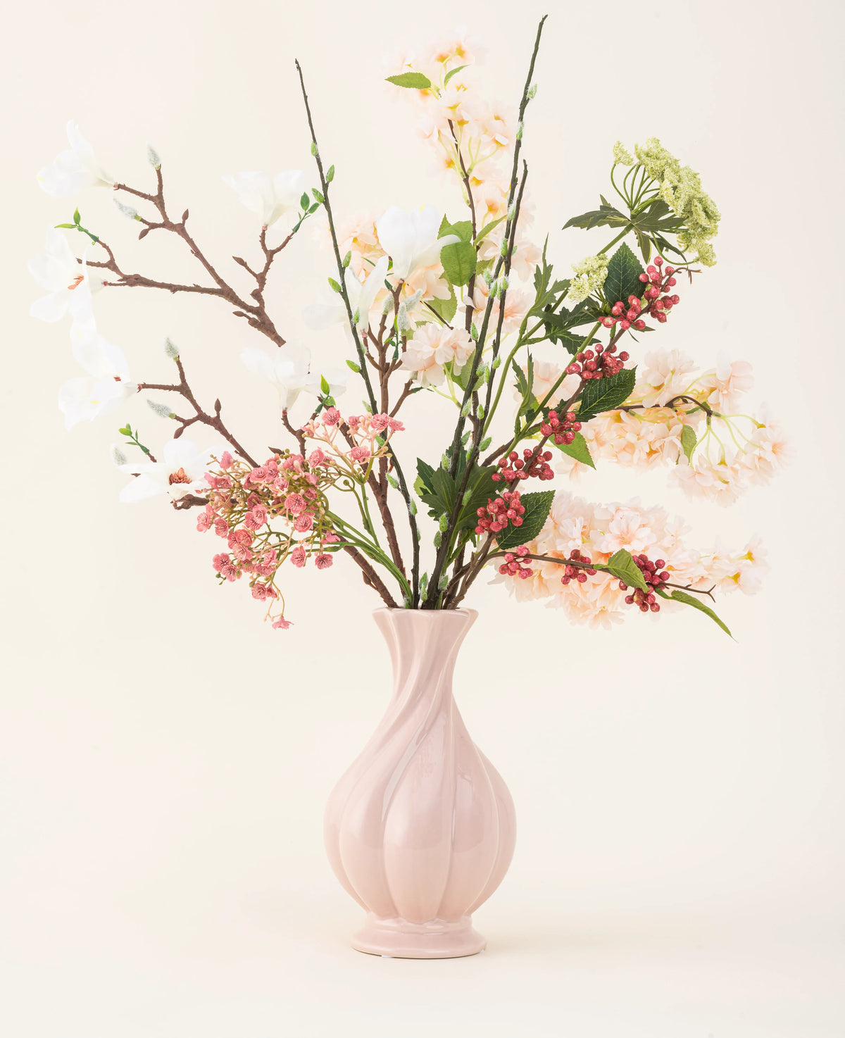 Un vase en céramique rose contient le bouquet de fleurs artificielles en soie Springtime Poem, avec des fleurs blanches, roses et vertes avec des feuilles vertes et des tiges brunes, sur un fond clair.