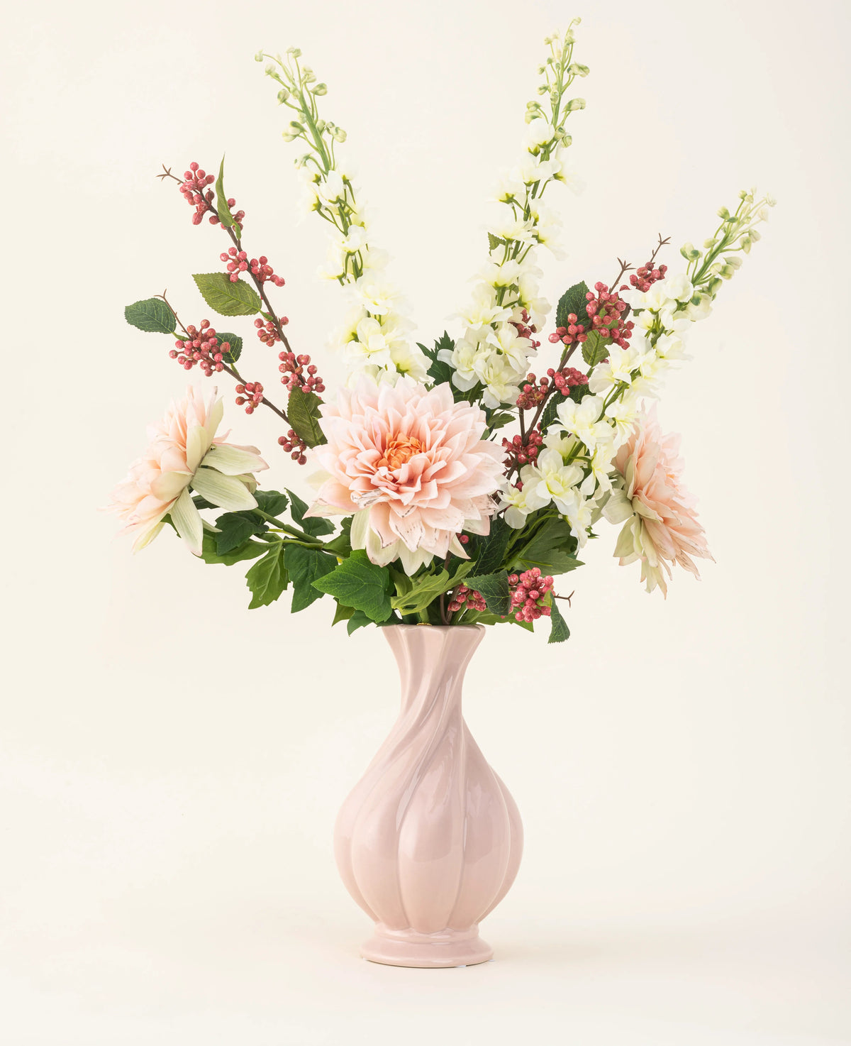 Un vase en céramique rose contient le bouquet de fleurs artificielles en soie Morning Whisper, avec des dahlias rose pâle, des mufliers blancs, des feuilles vertes et des grappes de petites baies rouges, sur un fond uni blanc cassé.
