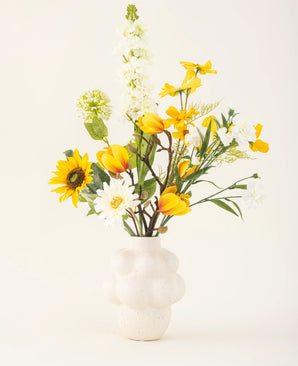 Le bouquet de fleurs artificielles en soie Sun Glow avec des tulipes jaunes, des tournesols, des marguerites et des feuilles vertes est placé dans un vase blanc structuré sur un fond uni blanc cassé.