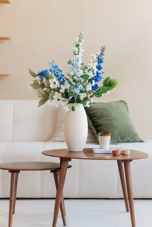 Un bouquet de fleurs artificielles en soie Cool Breeze composé de fleurs bleues et blanches se trouve dans un vase blanc sur une table basse ronde en bois avec une tasse, des bougies et un livre. Derrière se trouve un canapé blanc avec des coussins verts et beiges.