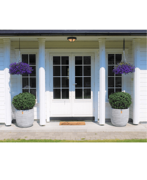 Une double porte blanche avec panneaux de verre est encadrée de deux Buis Artificiels sur tige UV d60 cm et de paniers suspendus à fleurs violettes, contre une maison blanche avec piliers et un petit tapis d'accueil à l'entrée.