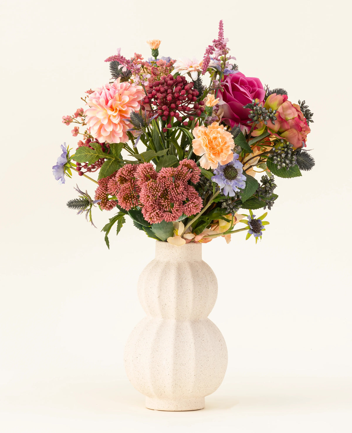 Dans un vase en céramique blanc incurvé se trouve le bouquet de fleurs artificielles en soie Pretty Pastel - un mélange coloré de fleurs durables telles que des roses roses, des dahlias, des fleurs violettes et oranges et de la verdure - magnifiquement disposées sur un fond clair.