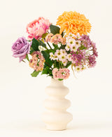 Un vase en céramique ondulée blanche présente le bouquet de fleurs artificielles en soie Peach Delight, comprenant un dahlia jaune, une pivoine rose, une rose violette et plusieurs petites fleurs blanches et roses sur un fond clair simple.