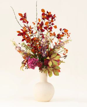 Le bouquet de fleurs artificielles en soie Golden Autumn présente un mélange artistique de feuilles d'automne et de fleurs rouges, oranges et violettes dans un vase en céramique blanche, sur un fond clair.