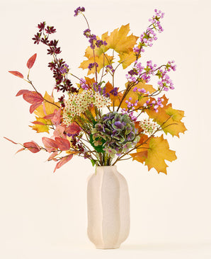 Le bouquet de fleurs artificielles en soie 'Wild Amethyst', avec des feuilles d'automne, des fleurs violettes et blanches et des hortensias verts, est présenté dans un vase en céramique blanche sur un fond uni et clair.