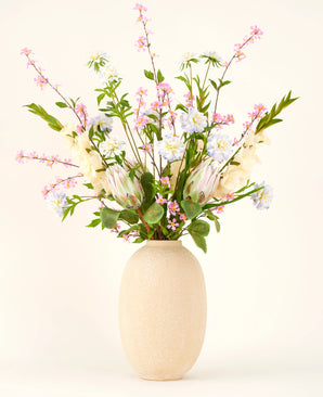Dans un vase à structure beige se trouve le bouquet de fleurs artificielles en soie Lots of Love XL, un bouquet noué à la main de fleurs en soie roses, blanches et bleues avec des feuilles vertes et de longues tiges sur un fond clair.