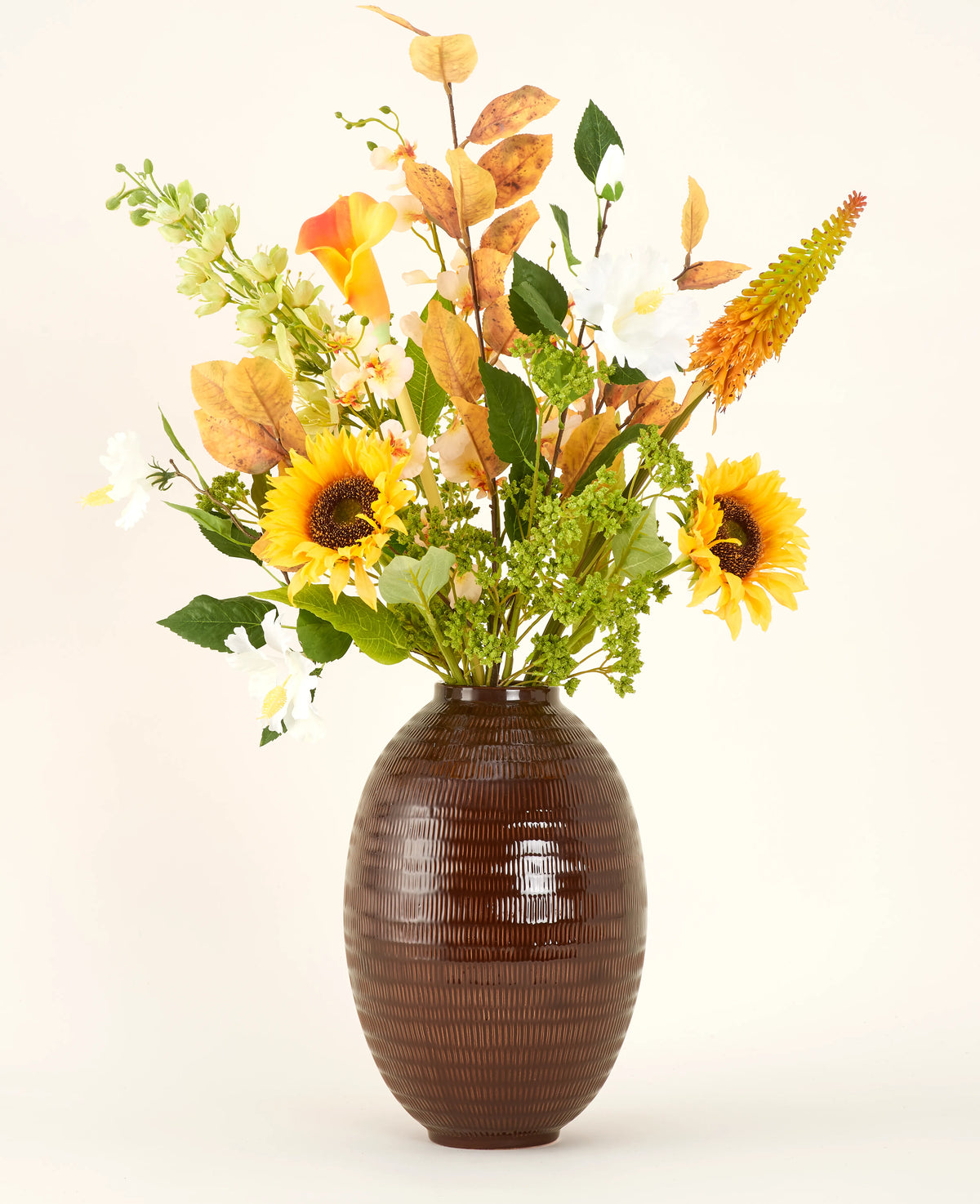 Un vase texturé marron montre le bouquet de fleurs artificielles en soie Amber Light, avec des tournesols, des fleurs blanches et oranges et un feuillage vert sur un fond uni.