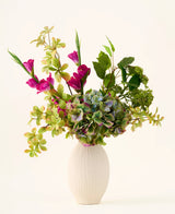 Un vase blanc texturé contient un bouquet d'hortensias verts, de branches feuillues et de fleurs en soie magenta vif sur un fond clair uni, créant un superbe bouquet « Cottage Garden » de fleurs artificielles en soie qui ajoutera un charme durable à n'importe quel espace.
