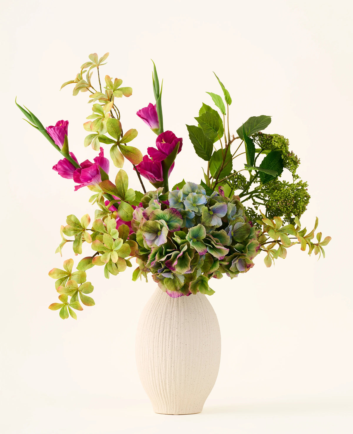 Un vase blanc texturé contient un bouquet d'hortensias verts, de branches feuillues et de fleurs en soie magenta vif sur un fond clair uni, créant un superbe bouquet « Cottage Garden » de fleurs artificielles en soie qui ajoutera un charme durable à n'importe quel espace.