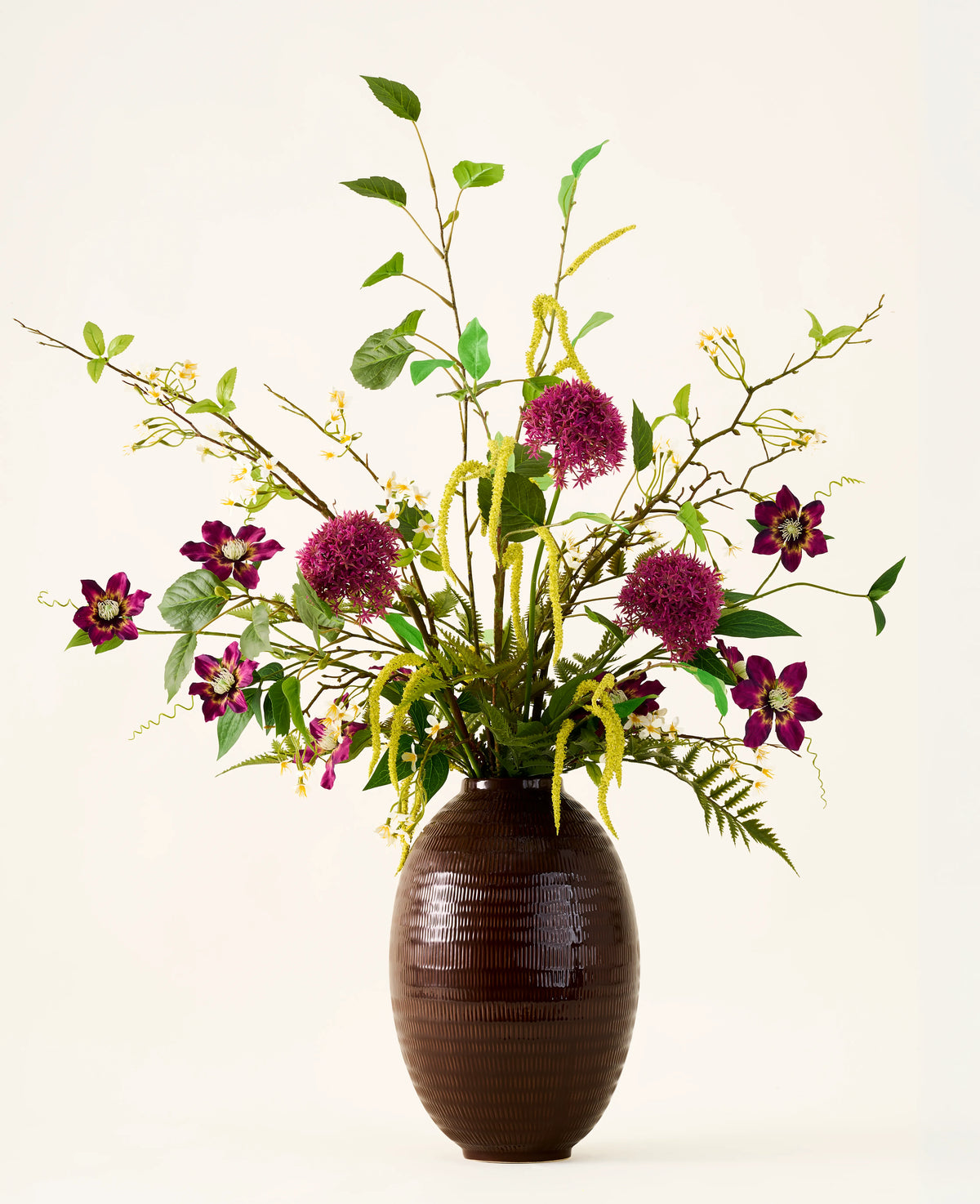 Dans un vase marron nervuré se trouve le bouquet "Woodland Charm" de fleurs artificielles en soie, avec des alliums violets, des fleurs rose foncé, des brindilles jaunes et des feuilles vertes sur un fond uni et clair.