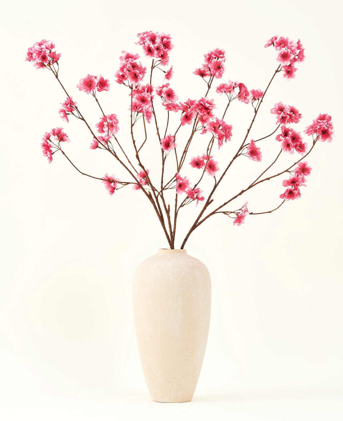 Le bouquet de fleurs artificielles en soie Fleur de cerisier fuchsia 114 cm ajoute une touche de couleur vibrante à votre intérieur avec ses branches saisissantes et ses fleurs fuchsia, joliment présentées dans un vase en céramique beige sur un fond uni blanc cassé.