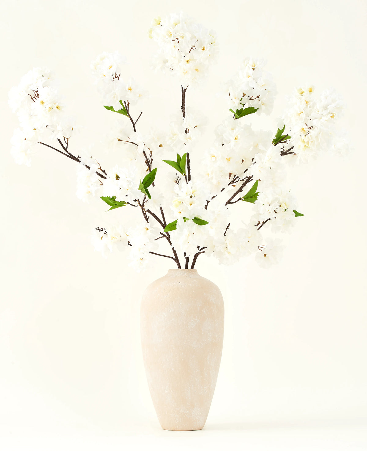 Le bouquet de fleurs artificielles en soie Fleur de cerisier crème 119 cm présente d'élégantes branches marron avec de fausses fleurs blanches douces et des feuilles vertes, présentées dans un simple vase beige sur un fond clair uni.
