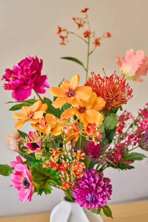Un bouquet vibrant de fleurs artificielles de Zijden, « Magnus di Lambo », composé d'un mélange de fleurs artificielles orange, roses et magenta, est disposé dans un vase blanc sur un fond neutre.