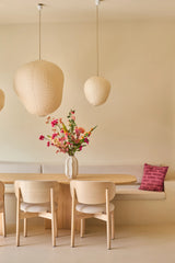 Une salle à manger minimaliste avec une table et des chaises en bois clair, des murs beiges, trois lampes suspendues en forme de lanternes en papier, un vase avec le bouquet de fleurs artificielles en soie "Wild Peach" qui apporte une touche de couleur vive et un canapé intégré avec un coussin décoratif rose sur le dessus.