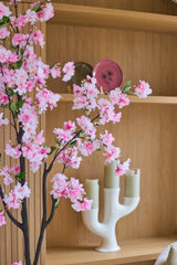 Une branche de fleur de cerisier artificielle 120 cm rose se dresse devant des planches de bois sur lesquelles reposent une assiette rouge, un bol et un bougeoir blanc à trois bras et des bougies vertes.