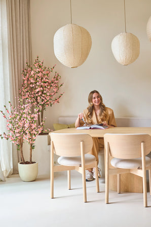 Une femme est assise, souriante, à une table avec un livre ouvert. La pièce est décorée avec des meubles en bois clair, des lanternes en papier, une douce lumière naturelle pénétrant à travers de hauts rideaux et une plante de cerisier artificielle de 120 cm rose qui ajoute une touche de fleur rose.