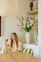 Une femme en costume crème est assise sur un banc avec une tasse à la main et un sourire aux lèvres. Derrière elle se trouve un grand vase avec le bouquet de fleurs artificielles en soie « Lots of Love XL », composé de fleurs roses, blanches et jaunes, avec des étagères en arrière-plan sur lesquelles sont exposés des objets décoratifs. Le cadre semble cosy et moderne.