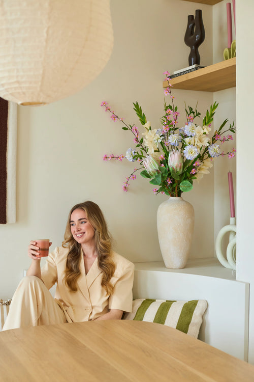 Une femme en costume crème est assise sur un banc avec une tasse à la main et un sourire aux lèvres. Derrière elle se trouve un grand vase avec le bouquet de fleurs artificielles en soie « Lots of Love XL », composé de fleurs roses, blanches et jaunes, avec des étagères en arrière-plan sur lesquelles sont exposés des objets décoratifs. Le cadre semble cosy et moderne.