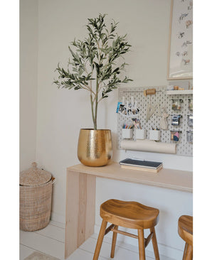 Un espace de travail moderne avec un bureau en bois, deux tabourets, une grande jardinière dorée avec un olivier artificiel de 120 cm pour une ambiance méditerranéenne, une planche à laver avec des fournitures de bureau et un panier en osier au sol comme détail décoratif.