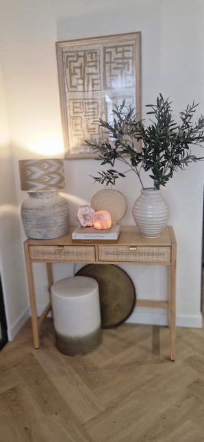 Une console en bois avec deux tiroirs contient une lampe, un grand vase blanc avec feuille d'olivier artificielle de 90 cm ignifuge, un plus petit vase, des livres et des pierres décoratives. Des accents méditerranéens et des œuvres d’art abstraites encadrées complètent le look ci-dessus. En bas se trouvent un tabouret et un plateau rond.