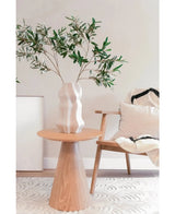 Dans un espace de vie moderne et minimaliste au décor méditerranéen, un vase blanc sculpté avec la feuille d'olivier artificielle 90 cm ignifuge trône sur une table ronde en bois, à côté d'une chaise en bois clair aux coussins blancs et noirs.