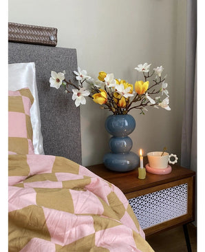 Une table de chevet avec un vase bleu contenant la Fleur Artificielle Magnolia 50 cm jaune/orange, une tasse à thé rose sur une soucoupe en forme de marguerite et une bougie allumée se dresse à côté d'un lit recouvert d'une couverture rose et marron aux motifs géométriques.