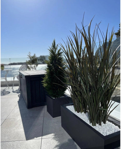 Le gazon artificiel Bordeaux UV 120cm en jardinières rectangulaires avec pierres blanches agrémente une terrasse extérieure moderne sur fond de ciel bleu et d'arbres au loin.