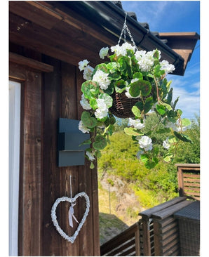 Le Bégonia Artificiel Blanc en Panier UV 70cm est accroché sur un mur en bois, avec un cœur blanc décoratif en dessous. De la verdure, une terrasse en bois et un ciel bleu partiellement nuageux complètent le décor.