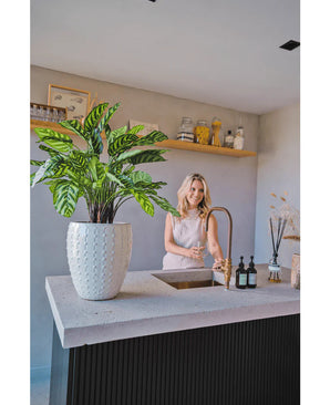 Une femme sourit devant un comptoir de cuisine moderne avec sa main sous le robinet. La Plante Artificielle Calathea Makoyana Deluxe UV 75 cm est au premier plan sur le comptoir. Des étagères ouvertes présentent des bocaux et des objets décoratifs en arrière-plan.
