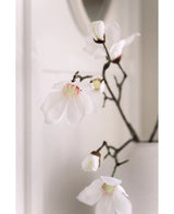 Un gros plan de la branche de magnolia artificielle Real Touch White 98 cm montre de délicates fleurs de magnolia blanches avec des cœurs roses sur des branches sombres dans un vase blanc, sur un fond clair légèrement décoloré.