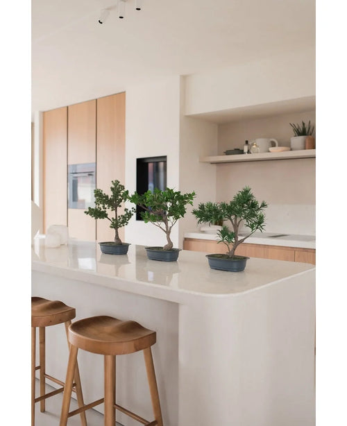Une plante artificielle Bonsai Juniper de 40 cm se dresse sur un îlot de cuisine blanc avec deux tabourets en bois, entourés d'armoires en bois clair, de plans de travail blancs et d'un décor minimaliste.