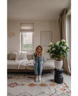 Une femme aux cheveux blonds bouclés est assise sur un canapé beige dans un salon lumineux et pose son menton sur ses mains. A côté d'elle se trouve la plante UV d'hortensia artificielle de 85 cm de haut, éclairée par la lumière à travers une fenêtre aux rideaux beiges.