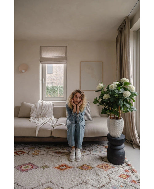 Une femme aux cheveux blonds bouclés est assise sur un canapé beige dans un salon lumineux et pose son menton sur ses mains. A côté d'elle se trouve la plante UV d'hortensia artificielle de 85 cm de haut, éclairée par la lumière à travers une fenêtre aux rideaux beiges.