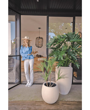 Une femme portant un chapeau, un chemisier bleu et un pantalon blanc s'appuie contre une porte vitrée. Au premier plan se trouve la plante artificielle Philodendron 125 cm. À l'intérieur, vous pouvez voir un espace de vie confortable et moderne.