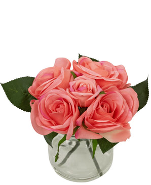 Le bouquet de roses artificielles Real Touch Pink 18 cm comprend cinq roses artificielles roses aux feuilles vertes, rapprochées les unes des autres dans un petit vase en verre rempli d'eau, sur un fond blanc.