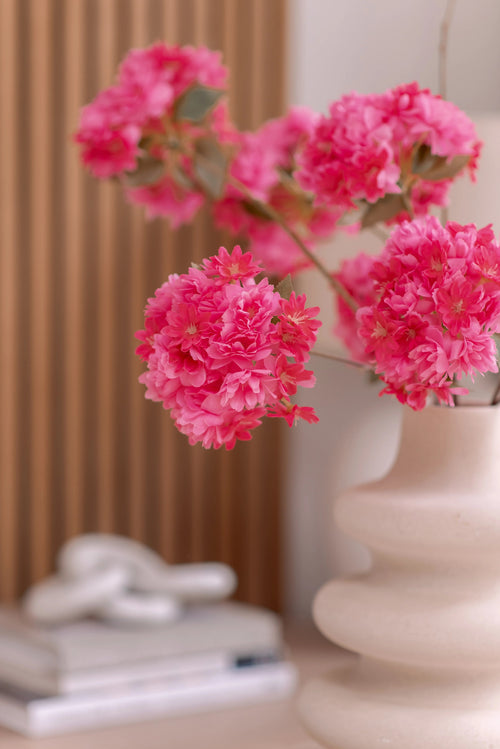 Un vase en céramique blanche contient une branche de fleur de cerisier artificielle de 85 cm rose. Le fond montre des panneaux de bois verticaux, une pile de livres et un petit objet décoratif au-dessus.