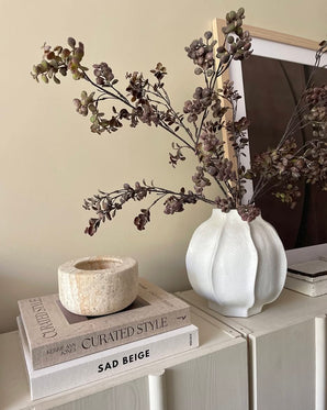 Sur une armoire lumineuse se trouve un vase blanc rempli de branche artificielle d'eucalyptus de 110 cm marron, à côté d'une pile de livres et d'un bougeoir en pierre, avec une photo encadrée en arrière-plan.
