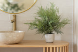 La Plante Artificielle Asperge 43 cm en pot décoratif se dresse sur un plan de travail en bois à côté d'un évier rond en pierre et d'un robinet doré, avec un miroir rond reflétant la scène sur un mur carrelé blanc.