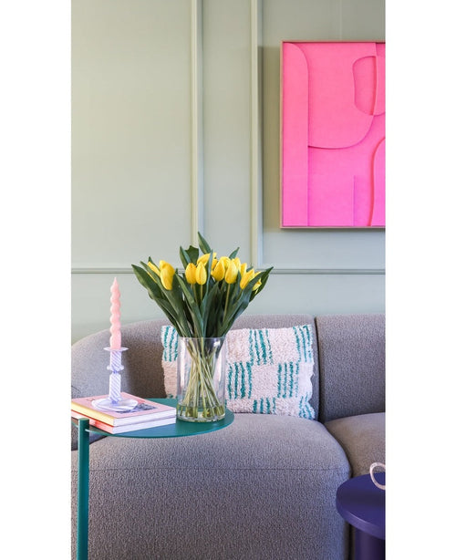 Le bouquet artificiel en soie Tulipes dans un vase Jaune se trouve sur une table d'appoint verte à côté d'une bougie torsadée rose, de deux livres et d'un canapé gris structuré avec un coussin à motifs. Une œuvre d’art abstraite rose vif est accrochée au mur vert clair.