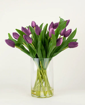 Le bouquet artificiel en soie Tulipes dans un vase Violet contient des tulipes artificielles violettes à longue tige avec des feuilles vertes luxuriantes, joliment disposées dans un vase en verre transparent sur un fond blanc uni.