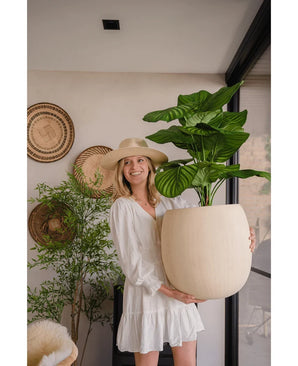 Une femme vêtue d'une robe blanche et d'un chapeau sourit alors qu'elle tient la plante artificielle Calathea UV 72 cm à l'intérieur, avec des paniers tressés et une autre plante en arrière-plan.