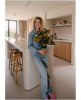 Une personne souriante vêtue de vêtements en jean se tient dans une cuisine moderne et tient une plante de magnolia artificielle dans un pot de 46 cm, une plante facile d'entretien. La cuisine, parfaite pour la décoration intérieure, comprend des armoires en bois, un îlot de cuisine blanc et deux tabourets de bar.