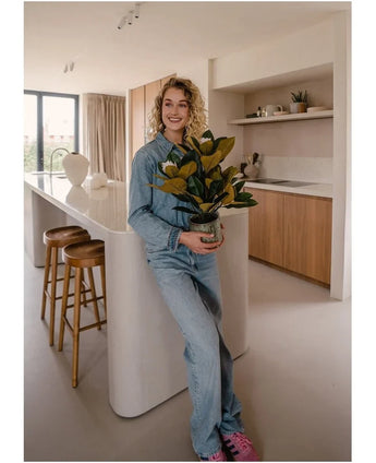 Une personne souriante vêtue de vêtements en jean se tient dans une cuisine moderne et tient une plante de magnolia artificielle dans un pot de 46 cm, une plante facile d'entretien. La cuisine, parfaite pour la décoration intérieure, comprend des armoires en bois, un îlot de cuisine blanc et deux tabourets de bar.