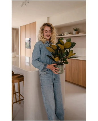 Une femme aux cheveux blonds bouclés, vêtue de vêtements en jean, se tient souriante dans une cuisine moderne avec la plante artificielle Magnolia dans un pot de 46 cm dans ses mains, entourée d'armoires et d'étagères en bois – un choix idéal pour tous ceux qui recherchent des plantes nécessitant peu d'entretien pour rehausser leur intérieur élégant.
