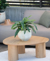 Une plante artificielle Bird's Deluxe de 60 cm se dresse sur une table basse ronde en bois devant un canapé beige, décoré d'un coussin vert et d'un chapeau marron suspendu au-dessus de l'accoudoir.