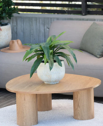 Une plante artificielle Bird's Deluxe de 60 cm se dresse sur une table basse ronde en bois devant un canapé beige, décoré d'un coussin vert et d'un chapeau marron suspendu au-dessus de l'accoudoir.