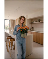 Une femme aux cheveux blonds bouclés, vêtue d'une chemise en jean et d'un jean, se tient dans une cuisine moderne avec dans ses mains une marguerite artificielle UV de 31 cm avec des fleurs orange vif. À côté d’elle se trouvent deux tabourets en bois près de l’îlot de cuisine.