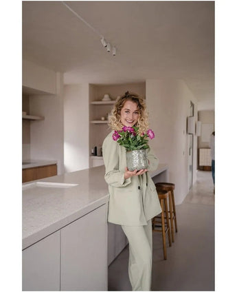 Een vrouw in een lichtgekleurd pak glimlacht terwijl ze een Kunstbloem Petunia UV 25 cm donkerroze vasthoudt; ze staat in een moderne keuken met neutrale tinten en een minimalistische inrichting.